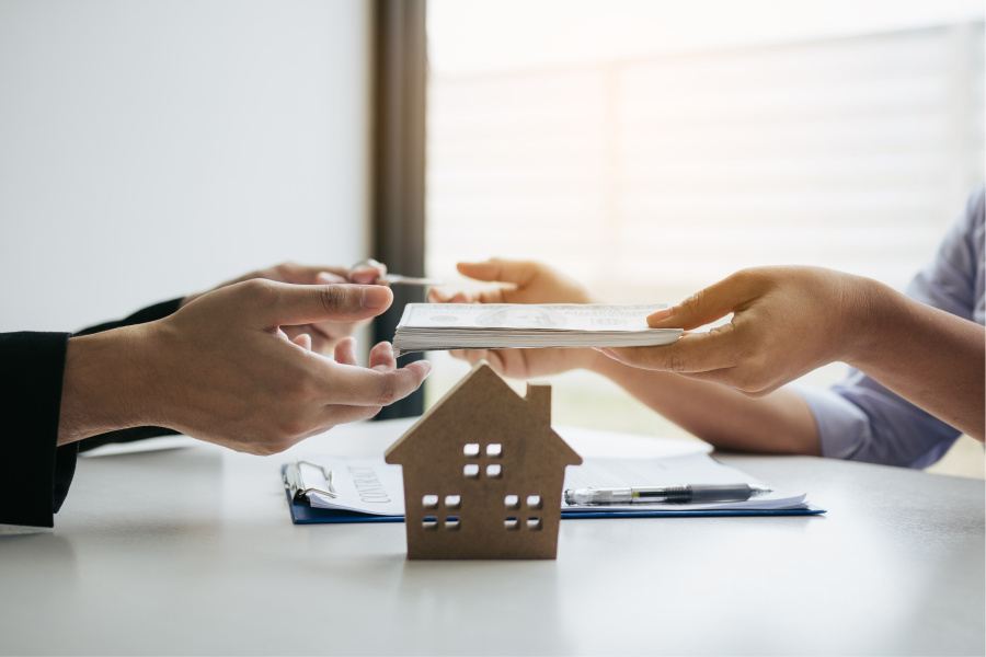 An image of a small wooden home block with the background of one professional hand handing a chunk of money to another professional hand